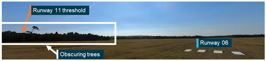 Photograph taken down runway 06 from the threshold. A highlighted line of trees obscured a portion of runway 11. The position of the obscured threshold of runway 11 is indicated with an arrow.